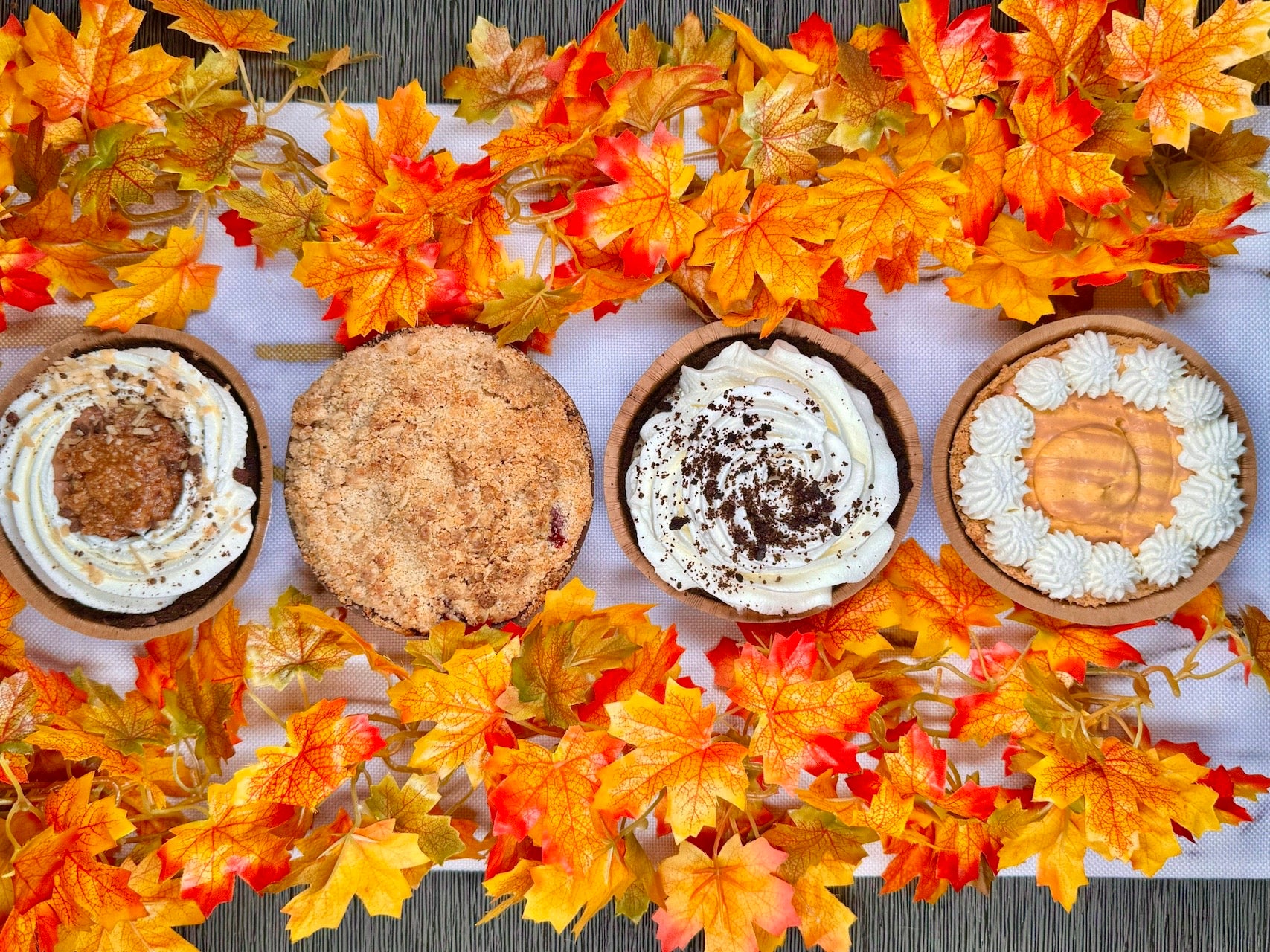 Four mini pies with whipped cream on a white surface surrounded by autumn leaves.